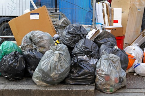 A collection of black and grey plastic rubbish bags filled with waste is piled on a paved sidewalk in an outdoor area. The bags vary in size, with some appearing larger and more tightly packed, while others are smaller and loosely tied. Among the waste bags, a brown cardboard box is partially visible on the left side, leaning against the bags. In the background, there are additional items including white and reddish containers, several cardboard boxes, and various packaging materials, suggesting a setting of rubbish clearance or waste disposal in a residential or commercial environment. The scene is illuminated by natural light, emphasizing the textures and contours of the bags and surrounding objects, with a slight industrial or urban atmosphere typical of waste removal operations carried out by companies like House Clearance Southfields, serving the Southfields area.