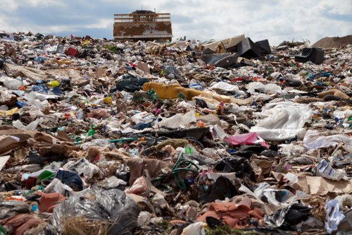 A large, open land area covered with a vast accumulation of mixed waste materials, including plastic bags, cardboard, paper, fabric, and various discarded items, creating a densely cluttered scene. In the background, there is a partially constructed or abandoned building, with an exposed framework and unfinished walls, situated under a partly cloudy sky. The scene suggests an ongoing or unresolved rubbish accumulation, typical of illegal dumping sites or unmanaged waste areas, which professional rubbish removal services like House Clearance Southfields aim to address. The ground appears uneven and heavily littered, with no visible vegetation, emphasizing the extent of waste contamination and the importance of thorough clearance efforts in the Southfields vicinity, postcodes SW18 or SW19.