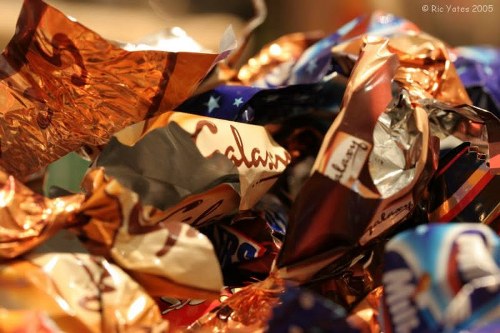 Close-up image of a tangled pile of crumpled, multicoloured foil and plastic wrappers, representing discarded confectionery packaging. The wrappers display various colours including copper, silver, blue, and red, with some featuring printed logos and text, although they are not clearly readable. The surface textures range from shiny and reflective to matte, with visible creases and folds from crumpling. The wrappers are stacked and intertwined, occupying the foreground of the image with no background context visible. This scene suggests waste materials typically found in household rubbish or post-gathering refuse, relating to rubbish removal services in Southfields. The lighting appears natural, highlighting the metallic sheen and vibrant colours of the wrappers, emphasizing their trash state. House Clearance Southfields handles the removal and disposal of such waste, often involving collection from residential areas, such as local Southfields properties, in postcode SW18, supporting efficient waste management practices in the area.